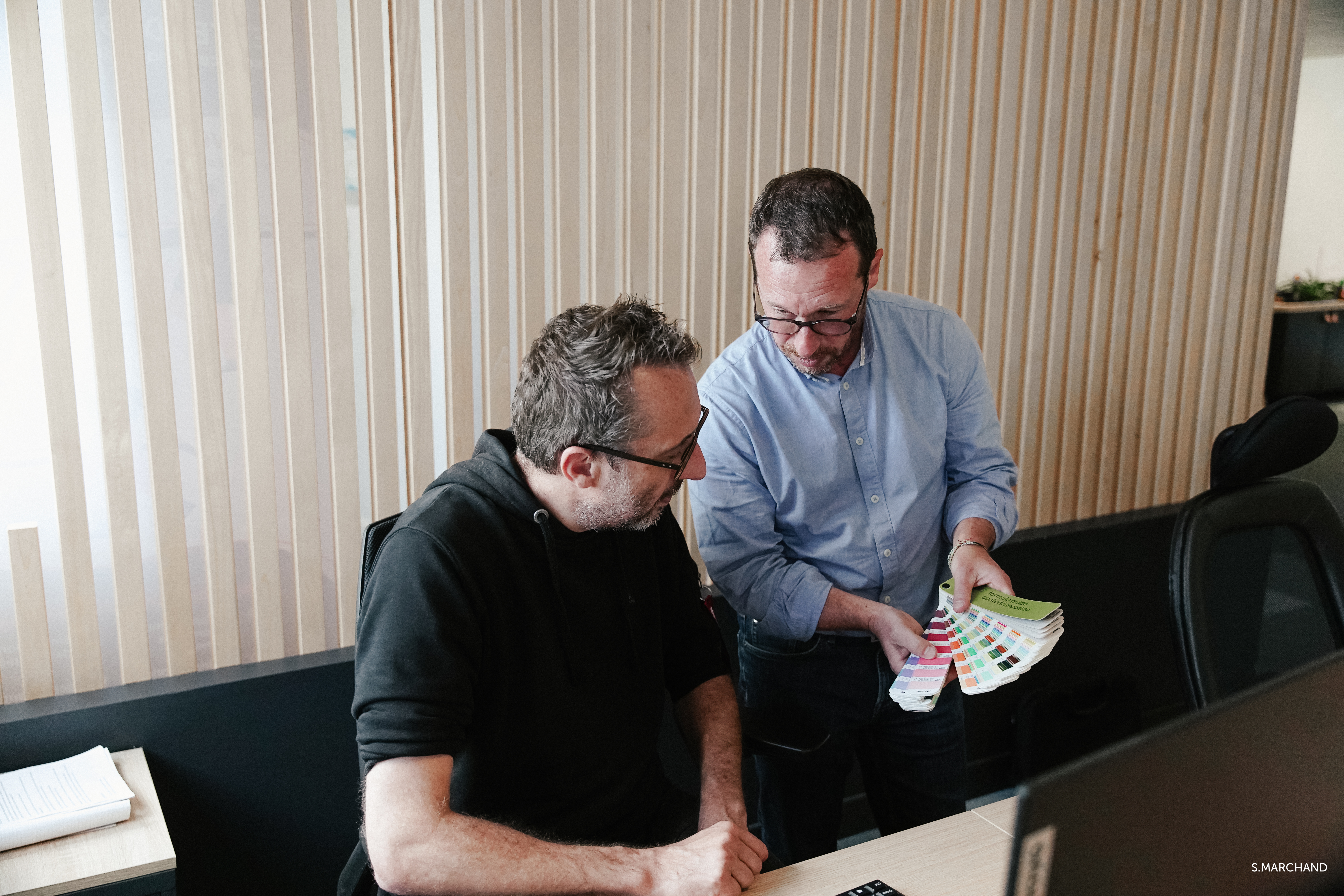 Deux hommes en discussion dans les bureaux de Nantes, l'un deux tient un référentiel de couleurs.